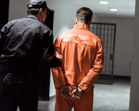 Rear View Of Prison Officer Leading Prisoner In Handcuffs In Cor